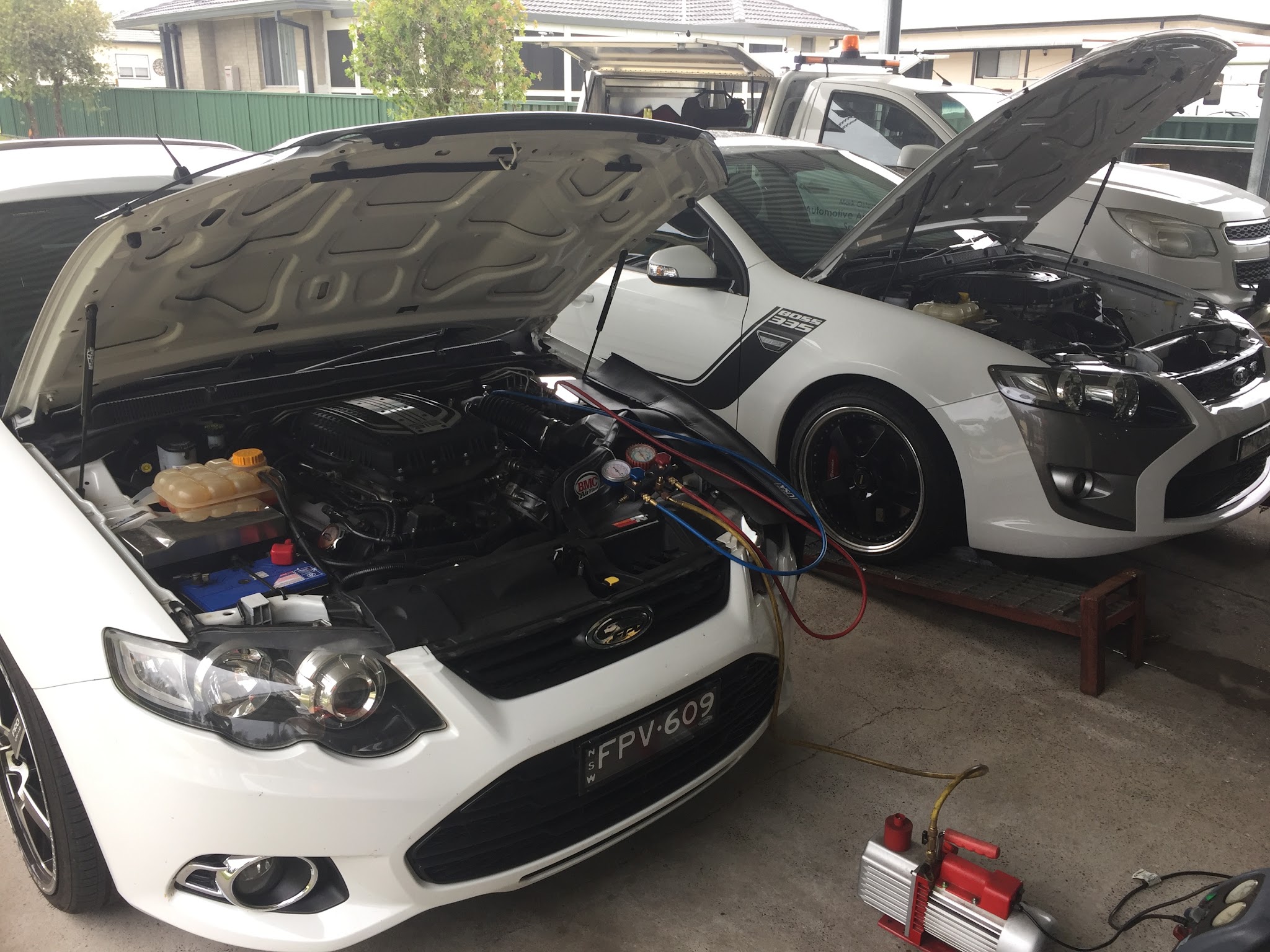Two white Ford Falcons under a carport with bonnets up, one connected to gauges and a running vacuum pump